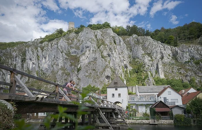 An der Balustrade der Holzbrücke vor Essing lehnt sich ein Pärchen an und schaut flussabwärts. Hinter  ihnen sieht man einige Häuser von Essing, dahinter die aufragenden Felswände.