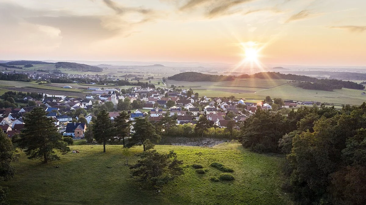 Der Blick von oben über grüne Wiesen hinab auf die Stadt Monheim. Die Sonne strahlt am Horizont und ist fast untergegangen.