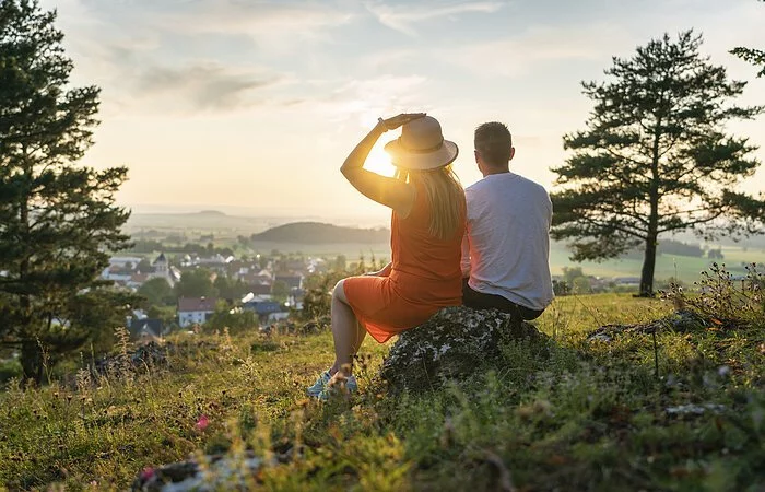 Ein Pärchen sitzt oberhalb von Monheim auf einem Stein umgeben von grüner Wiese und eingerahmt von zwei Bäumen. Die beiden genießen den Sonnenuntergang und den schönen Blick auf Monheim.