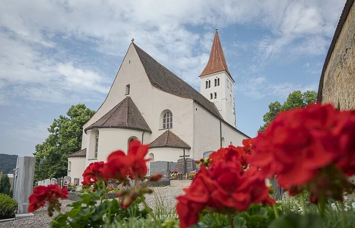 Die Basilika von außen. Rechts ist die Stadtmauer angedeutet und im Vordergrund blühen rote Blumen.