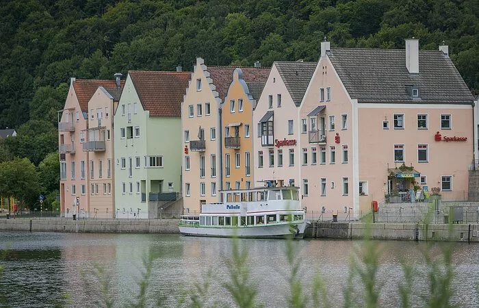 Der Main-Donau-Kanal im Vordergrund mit grünen Gräsern. Dahinter hat ein Personenschiff an der Schiffsanlegestelle Riedenburg angelegt. Die bekannte Promenade mit ihren Häusern ist ebenfalls zu sehen. Eingerahmt vom grün der Wälder.