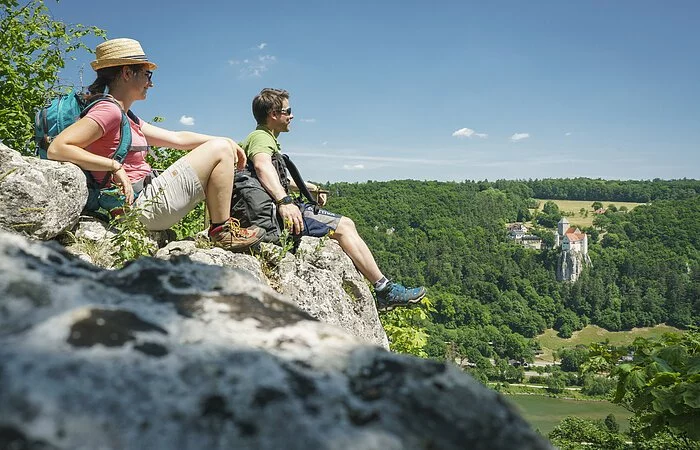 Auf dem Aussichtspunkt gegenüber Burg Prunn sitzen im Vordergrund ein Pärchen mit Blick ins Tal gerichtet.