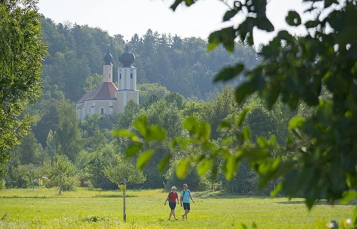 Ein Pärchen wandert unterhalb der Wallfahrtskirche Breitenbrunn entlang satter grüner Wiesen. Die Wallfahrtskirche ist von grünen Wäldern umgeben.