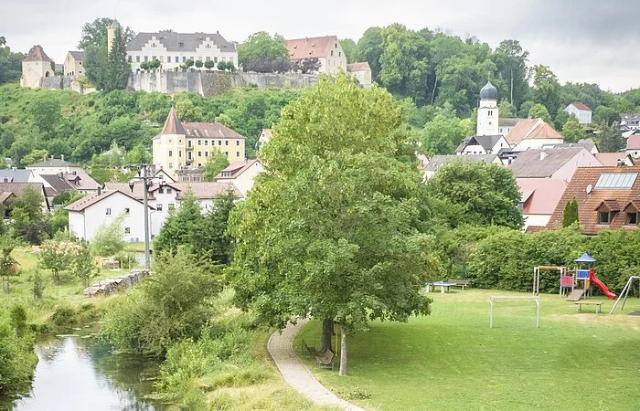 Drohnenaufnahme von Möhren. Im Vordergrund sind 2 Wanderer auf einer Holzbrücke über den Möhrenbach zu sehen. Im Hintergrund thront Schloss Möhren über dem Dorf.