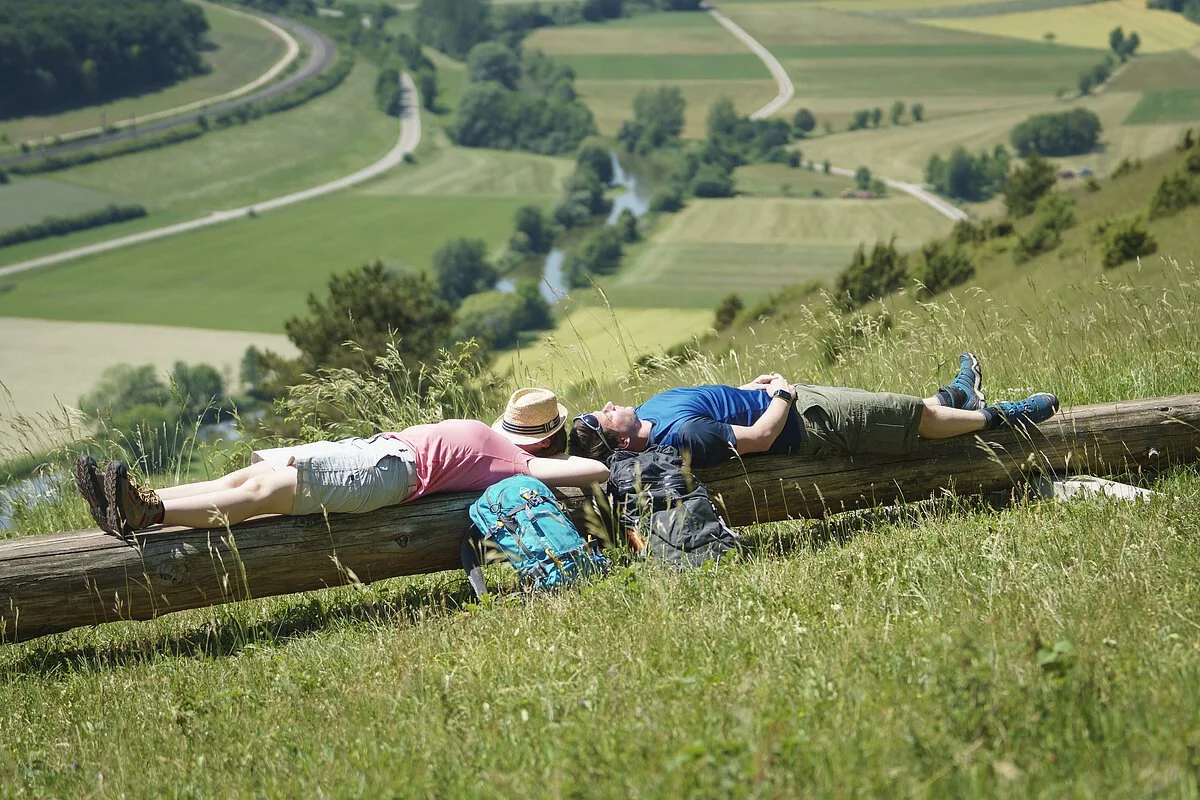 Auf der langen Bank liegen eine Frau links und ein Mann rechts. Ihre Rucksäcke an die Bank gelehnt. Hinter den beiden hat man einen guten Blick ins Tal.