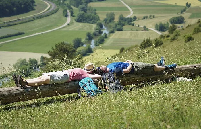 Auf der langen Bank liegen eine Frau links und ein Mann rechts. Ihre Rucksäcke an die Bank gelehnt. Hinter den beiden hat man einen guten Blick ins Tal.