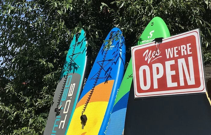 Bunte SUP-Boarde lehnen an einem Busch. Daran hängt ein Schild: Yes, we are open.