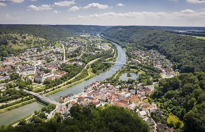 Der Blick auf Riedenburg von oben. Mitten durch die Stadt verläuft der Main-Donau-Kanal. Umrandet wird sie von grünen Wäldern. Der Himmel ist blau und einige Wolken sind zu sehen.