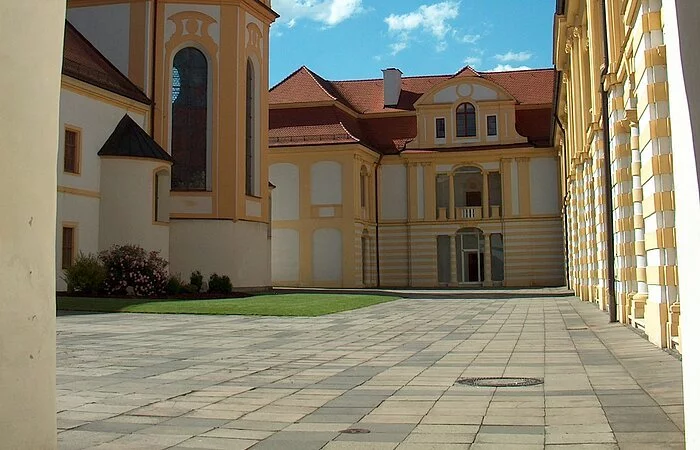 Ein gemauerter Torbogen umrandet das Bild, durch den man in den Innenhof gelangt. Der Hof zeichnet sich durch Grünflächen und den hellgepflasterten Boden aus. Um ihn herum stehen mehrere Gebäude. Sie sind in weiß und gelb gehalten. Auf der linken Seite erstreckt sich ein Teil des Kirchenschiffs. Auf der rechten Seite und im Hintergrund sind die Gebäude, die Räume der Maria-Ward Realschule beherbergen.