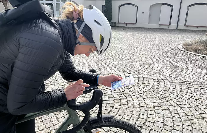 Eine Radlerin mit weißem Helm beugt sich zu ihrem Lenker auf dem sie das Heftchen aufgeschlage hat und mit einem Bleistift eine Notiz einträgt. Im Hintergrund ist ein Gebäude sichtbar.