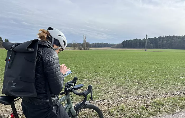 Eine Radlerin mit weißem Helm beugt sich zu ihrem Lenker auf dem sie das Heftchen aufgeschlagen hat und mit einem Bleistift eine Notiz einträgt. Im Hintergrund ist ein grünes Feld sichtbar.