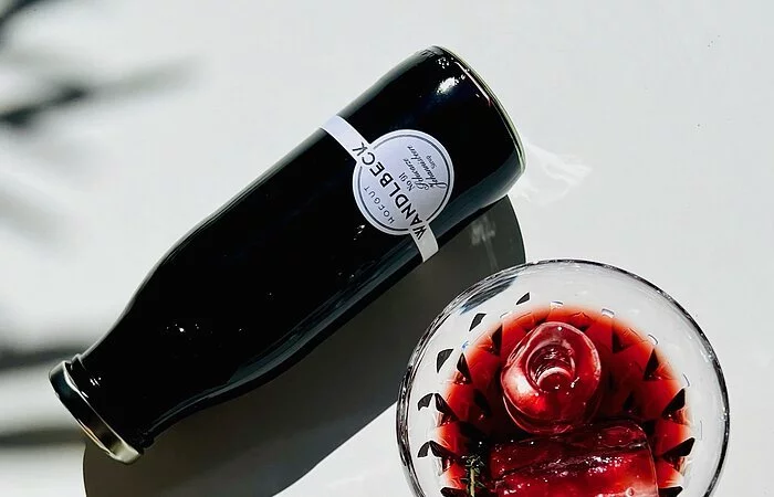 Auf einer Spiegelplatte steht ein Likörglas in dem ein roter Likör eingefüllt ist. Eine Kirsche liegt darin. Neben dem Glas liegt eine kleine Flasche mit rotem Inhalt und ein Stein mit einer Versteinerung.