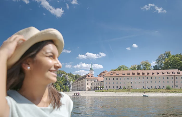 Eine junge Frau mit Strohhut und sommerlicher Kleidung sitzt in einem Ausflugsschiff mit grüner Sitzbank. Neben ihr liegt die Sonnenbrille auf der Bank. Im Hintergrund ist die Donau und das Kloster Weltenburg zu sehen. Am hellblauen Himmel sind einige weiße Schäfchenwolken zu sehen.
