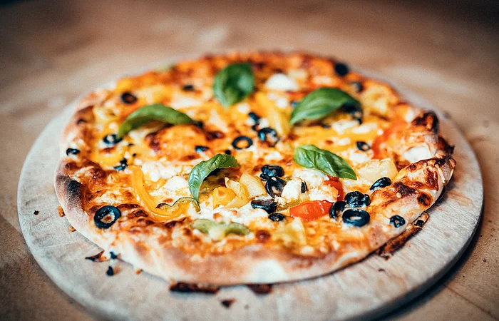 Eine Pizza mit fünf Basilikumblättern, Oliven und leckeren Käse liegt auf einem Holzbrett. Darunter sieht man das Holz eines Tisches.