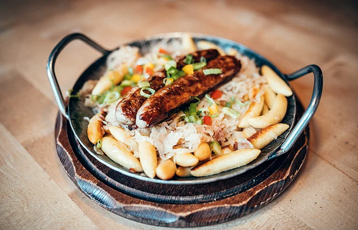 Zwei fränkische Bratwürste liegen wohl gebetet auf Schupfnudeln und Sauerkraut. Das Gericht ist in einem Eisenteller mit zwei Griffen angerichtet.