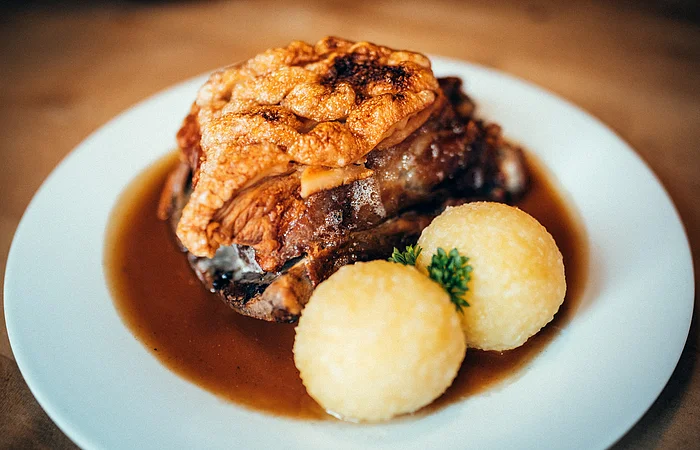 Auf einem weißen Teller sind zwei Kartoffelknödel mit Petersilie dazwischen angerichtet. Dahinter liegt das knusprige Schäuferl. Leckere Bratensoße zum Tunken füllt das weiße Teller aus.