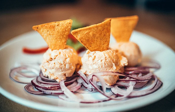 Drei Kugeln Obazda drapiert auf roten Zwiebeln. In jeder Kugel steckt ein Nacho. Im Hintergrund ist Paprika und Salat zu entdecken.