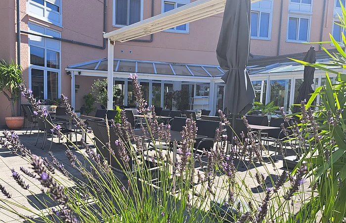 Der lila blühende Lavendel direkt an der Terrasse versperrt ein wenig die Sicht. Im Hintergrund ist die rosafarbende Fassade des Hotels erkennbar, sowie Sonnenschirme.