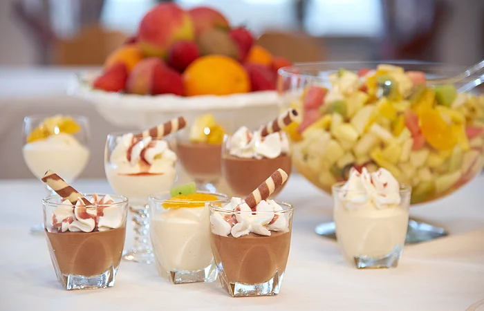 Lecker sechs Dessertgläser mit weißer und Schokoladenmousse stehen auf einem Tisch mit weißer Tischdecke. Im Hintergrund ist eine große Schale mit Obstsalat und eine Obstschale.