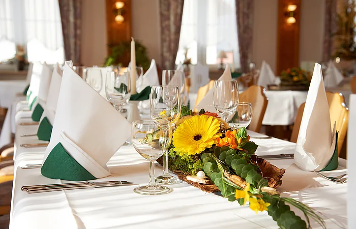 Das Restaurant ist mit langen Tischreihen bestückt. Die Tische sind festlich mit weißer Tischdecke und Servietten mit grün-weiß, sowie Gläsern gedeckt. Am Tischanfang steht eine schöne Blumendeko.