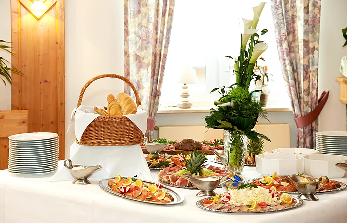 Es leckeres kaltes Buffet ist vor einem Fenster aufgebaut. Es gibt Platten mit Wurst, Lachs und Käse. Erhöht steht der große gut gefüllte Brotkorb. Eine Blumendeko und Teller gehören ebenfalls dazu.
