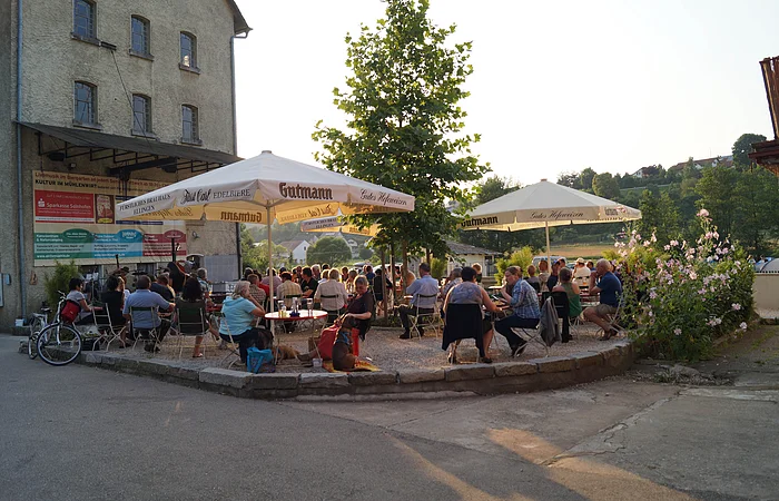 Der Biergarten mit weißen Sonnenschirmen ist gut besucht. Im Hintergrund steht ein einzelner Baum. Links ist ein Gebäude zu sehen. Die Sonne steht schon recht tief.