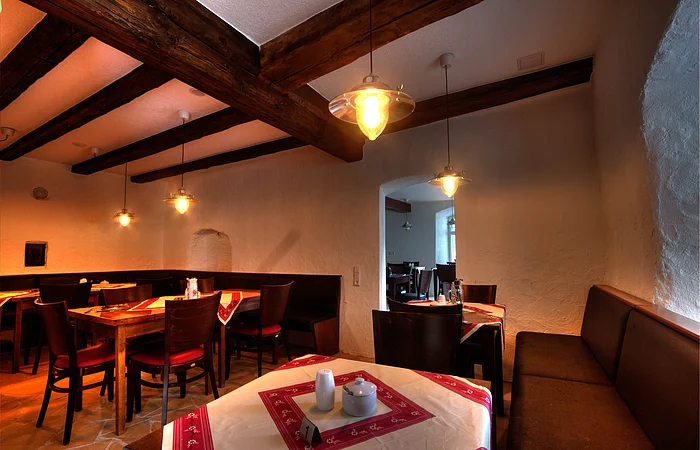 Der Gastraum ist mit dunklen Tischen und Stühlen eingerichtet. Die Tischdecken sind rot-weiß und die Deckenleuchten hängen von der Decke.