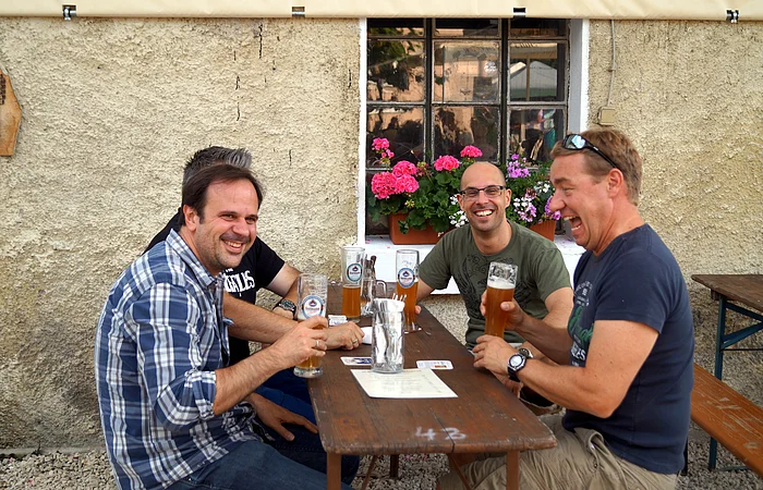 An einer Biertischgarnitur vor dem Wirtshaus sitzen vier Männer mit jeweils einem gefüllten Bierkrug in der Hand.