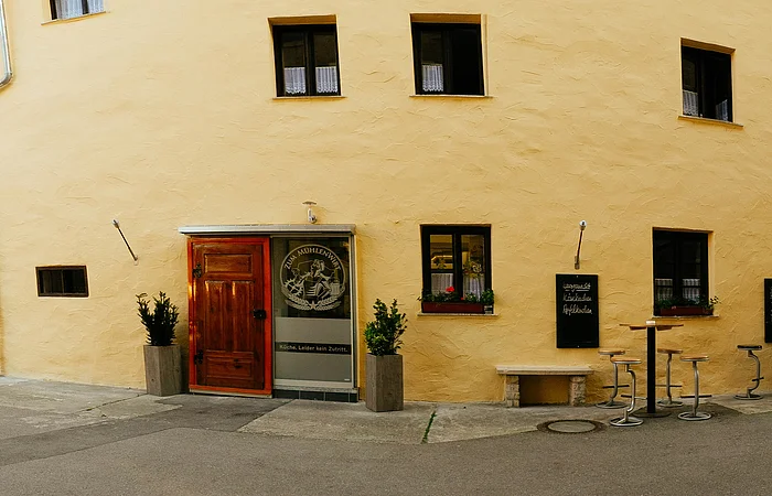 Das Wirtshaus von außen ist hellgelb gestrichen. Das Gebäude ist recht groß und rechts sind drei Bistrotische vor dem Haus aufgestellt.