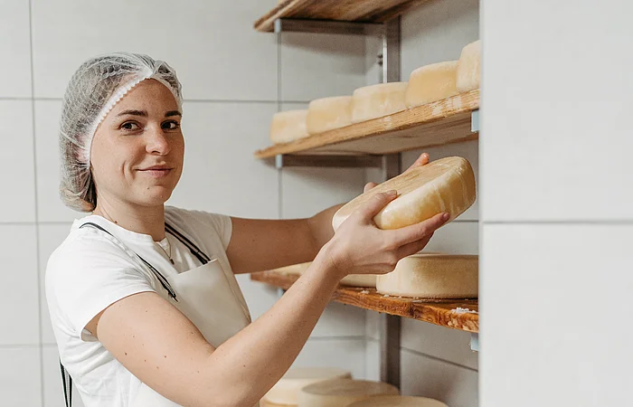 Eine weibliche Person ganz in weiß gekleidet mit  Haarnetz räumt in einem weiß gefliestem Raum große Käselaibe auf Holzregalbretter, die an der Wand montiert sind.