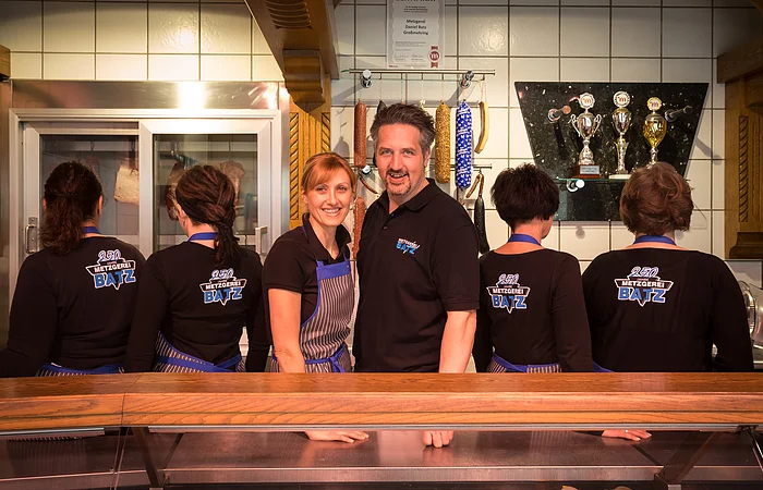 Hinter der Ladentheke hat sich das gesamte Team versammelt. Die Mitarbeiter sind mit dem Rücken zu sehen, auf ihren T-Shirts prangt das Logo der Metzgerei. In der Mitte ist das Ehepaar Batz zu sehen.
