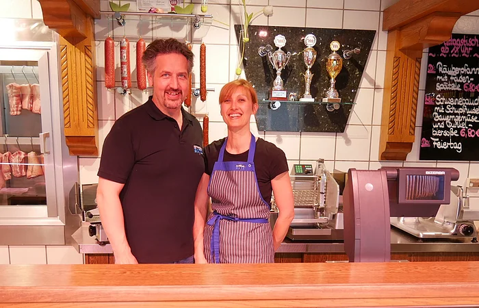 Das Ehepaar Batz ist hinter der Ladentheke zu sehen. Wurstprodukte und Pokale sind an der Hinterseite zu sehen.