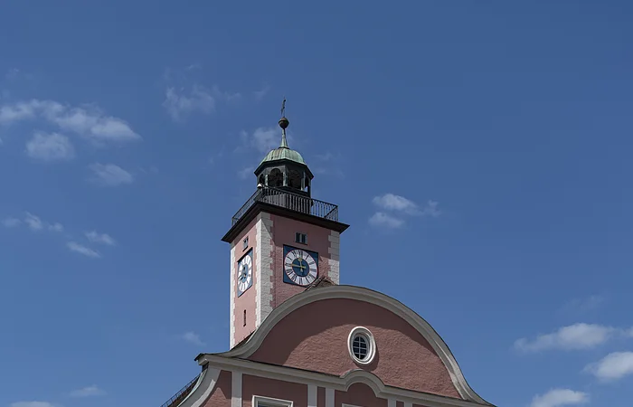 Das Bild zeigt ein prächtiges vierstöckiges Haus mit einem geschwundenen Giebel. Darüber thront im Hintergrund ein Glockenturm mit Ziffernblätter und einem Kupferdach sowie einem Balkon, der um den gesamten Turm führt. Das Rathaus und der Turm sind rotbraun gestrichen. Der Eingang in Form eines zweiflügeligen Holztors befindet sich in der Mitte der Vorderseite, direkt darüber ist ein Balkon zu erkennen, der durch zwei Säulen links und rechts des Eingangstores gestützt wird.