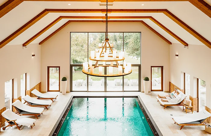 Ein Blick von oben herab auf den Swimmingpool. Links und rechts sind Liegen mit weißen Auflagen aufgereiht. Ein großes Fenster lässt viel Licht von außen herein.