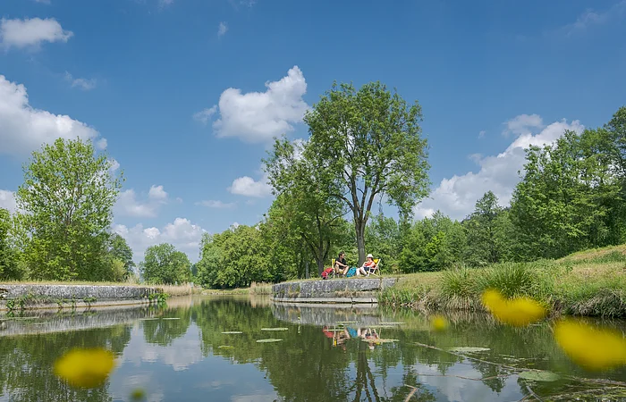 Am Ludwig-Donau-Main-Kanal der mittig fließt, haben es sich ein Pärchen am rechten Ufer in Liegestühlen gemütlich gemacht. Rechts und links stehen Bäume, der Himmel strahlt in blau.