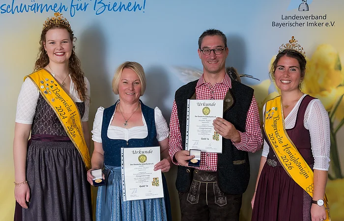 Vier Personen in Tracht stehen vor einer blauen Poster Wand. Drei Frauen und ein Mann mit. Die beiden Frauen links und rechts tragen eine gelbe Schärpe während die beiden innen stehenden Personen eine Urkunde vor sich halten.