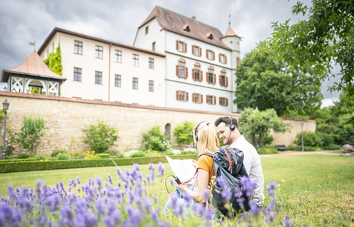 Auf der Wiese vor dem Stadtschloss sitzen 2 Touristen. Sie lauschen der Treuchtlinger Stadttour über die Kopfhörer der Audioguides.