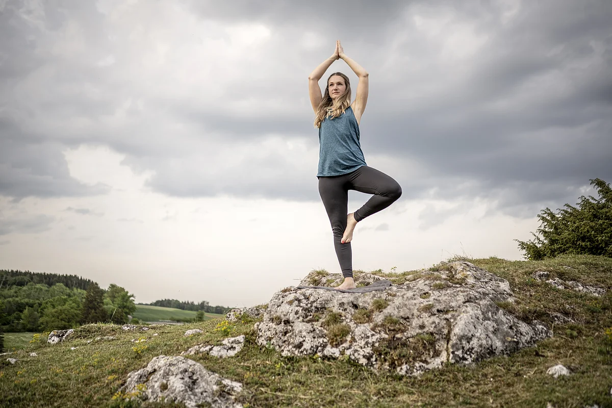 junge Frau steht in der Natur auf einem Felsen in einer einbeinigen Yogafigur mit nach oben gestreckte Armen.