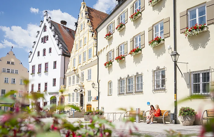 Zwei Freundinnen sitzen auf einer Bank vor dem Verwaltungsgebäude am historischen Marktplatz und unterhalten sich.
