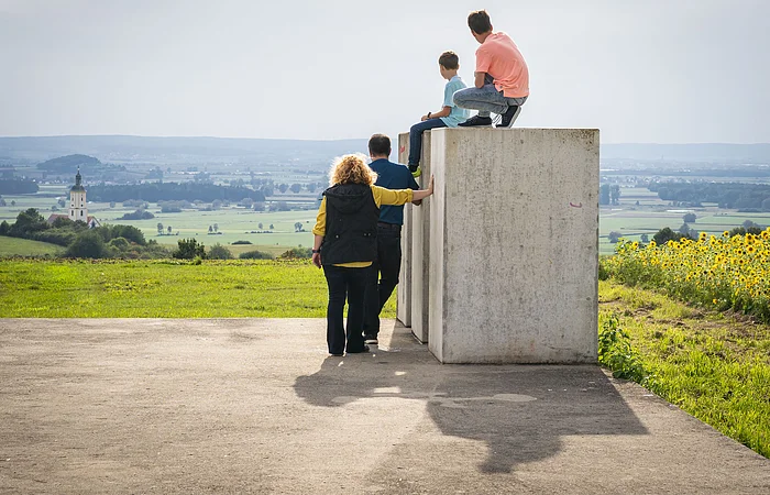 Die Kinder sitzen auf den Steinen der Zeitpyramide und die Eltern stehen daneben. Gemeinsam blicken sie in das Ries und genießen den tollen Ausblick.