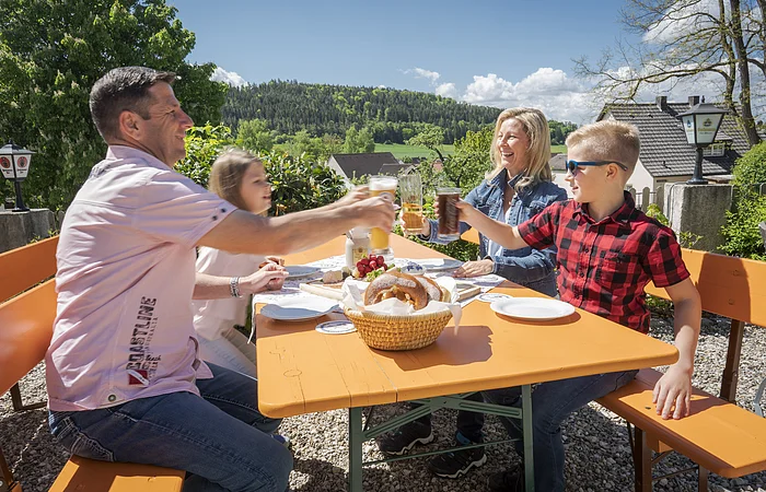 Biergarten, Nahaufnahme einer Familie mit zwei jüngeren Kindernv prosten sich bei einer Brotzeit mit Brezen, Käse und Radieschen zu.