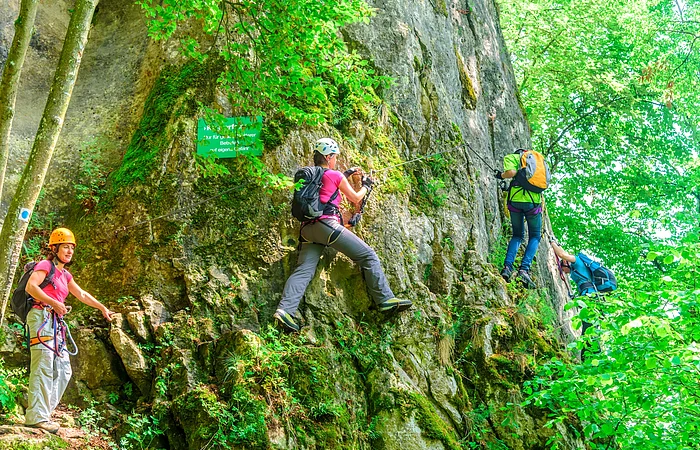 Vier Kletterer mit Helm und Ausrüstung sind am  Klettersteig unterwegs. Drei klettern bereits am Felsen entlang. Der Vierte steht noch und wartet, dass auch er an den Felsen kann.