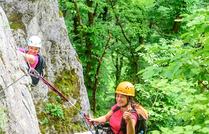 Eine Kletterin mit blonden Haaren hängt gesichert locker am Seil mitten am Felsen. Eine zweite ist schon ein Stück weiter und grinst herüber.