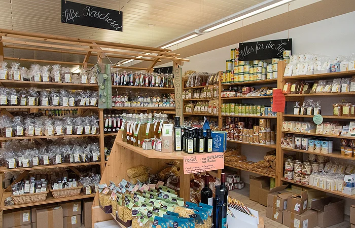 Blick in den Hofladen. Hohe Holzregale, gefüllt mit regionalen Produkten in Tüten, Dosen und Gläsern. Davor ein Tisch, auf dem weitere eingepackte Produkte präsentiert werden.