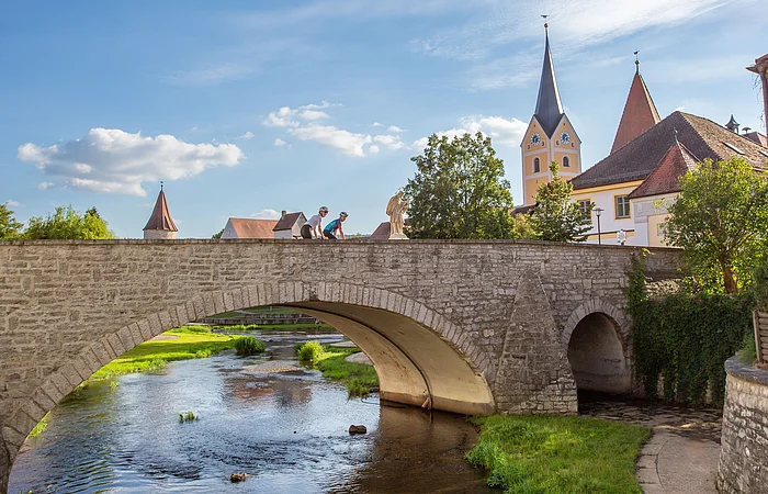 Zwei Renradfahrer fahren in Berching über eine Brücke und schauen dabei auf den Sulzpark und den Fluss