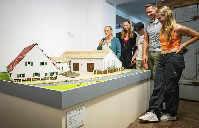 Vier Gäste in der Abteilung Möbel aus dem 17. Jahrhundert. Sie stehen auf der rechten Seite und blicken auf das Modell eines Jura-Bauernhofes.