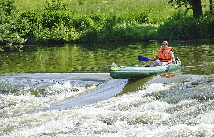 Ein Bootsfahrer mit oranger Rettungweste und Paddel steht mit seinem grünen Kanu am Anfang der Bootstreppe in der Altmühl. Das Wasser rauscht hier an dem steinernen Wehr hinunter. Die Bootsrutsche ist eine Vertiefung für die Fische und auch Boote. Das Ufer ist sattgrün auf dem die Sonne scheint.
