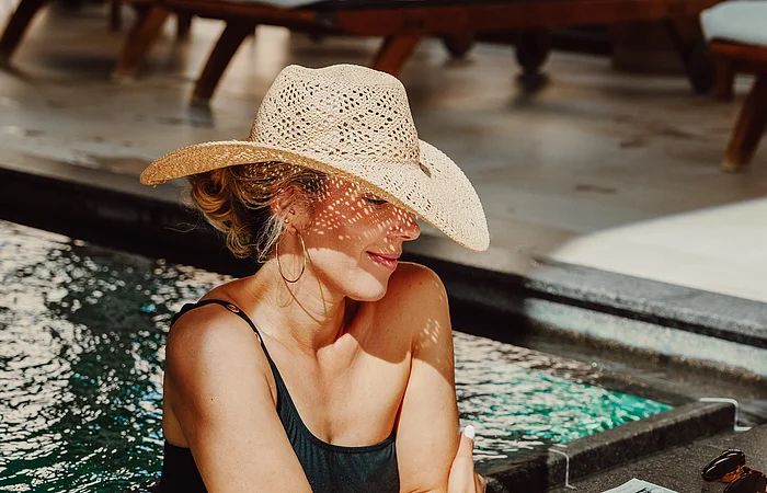 Eine Frau im schwarzen Badeanzug mit Strohhut kühlt sich im Außenpool ab. Sie lehnt am Beckenrand und liest Zeitung.