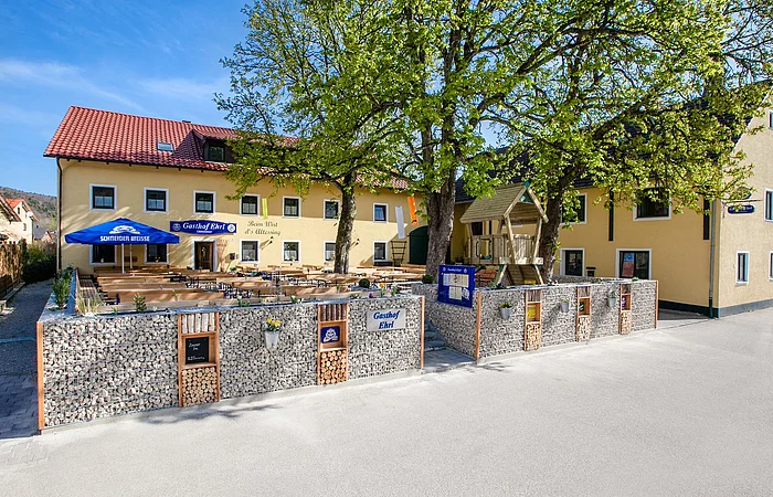 Außenansicht des Gasthofs. Im Biergarten davor sind die blauen Sonnenschirme offen und der Baum spendet den Gästen Schatten.