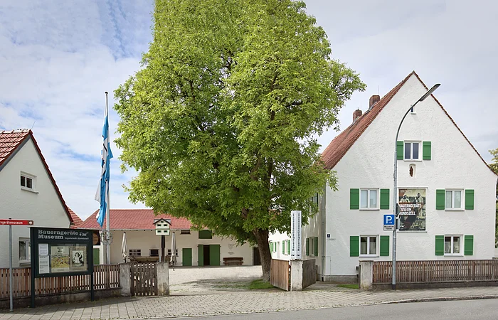 Der Eingangsbereich des Museums mit seinen Gebäudlichkeiten. Links ist zudem eine Infotafel und eine Fahne angebracht. Der Blick in den Hof zeigt ein weiteres Gebäude und einen großen Baum.